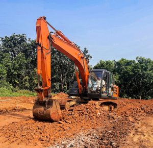 An orange excavator construction machine working on a road construction project. Plenty of red soil and crushed stone with clear blue sky on the background